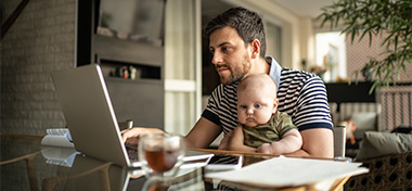 Man holding his newborn baby and working on laptop at home