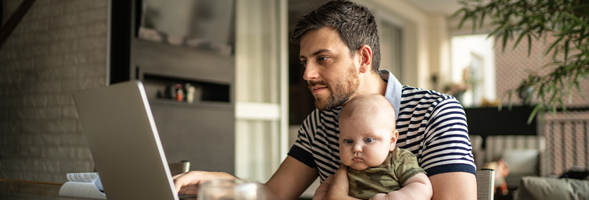 Man holding his newborn baby and working on laptop at home